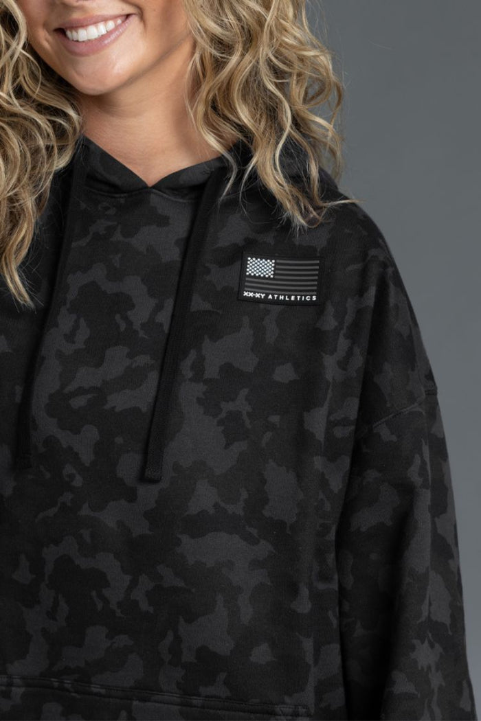 Person wearing a black camouflage hoodie with a logo on a gray background || Black Camo