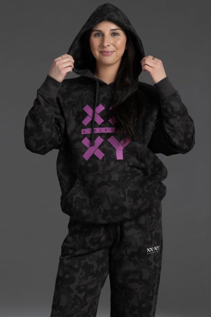 Person wearing a black hoodie with a purple logo on a gray background || Black Camo
