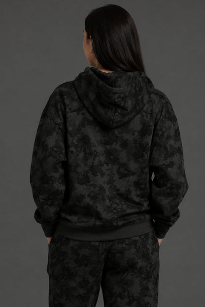 A person shown from the back wearing a black hooded sweatshirt and matching black patterned pants against a dark gray studio background. || Black Camo